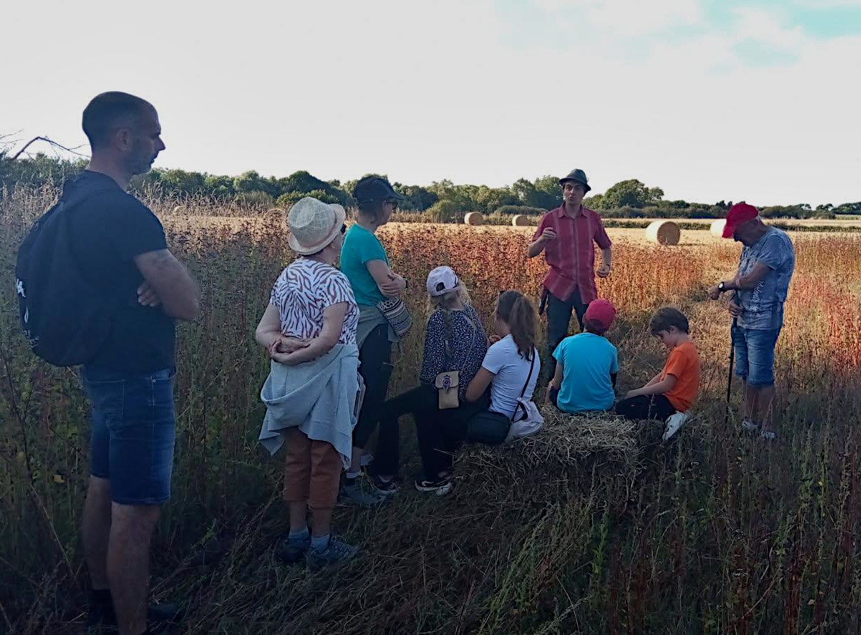 Un homme, chapeau gris, chemise rouge, parle à un groupe composé d'enfants et d'adultes. Ils sont dans un verger, on aperçoit des pommiers derrière.