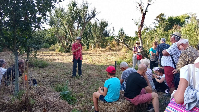 Un homme, chapeau gris, chemise rouge, parle à un groupe composé d'enfants et d'adultes. Ils sont dans un verger, on aperçoit des pommiers derrière.